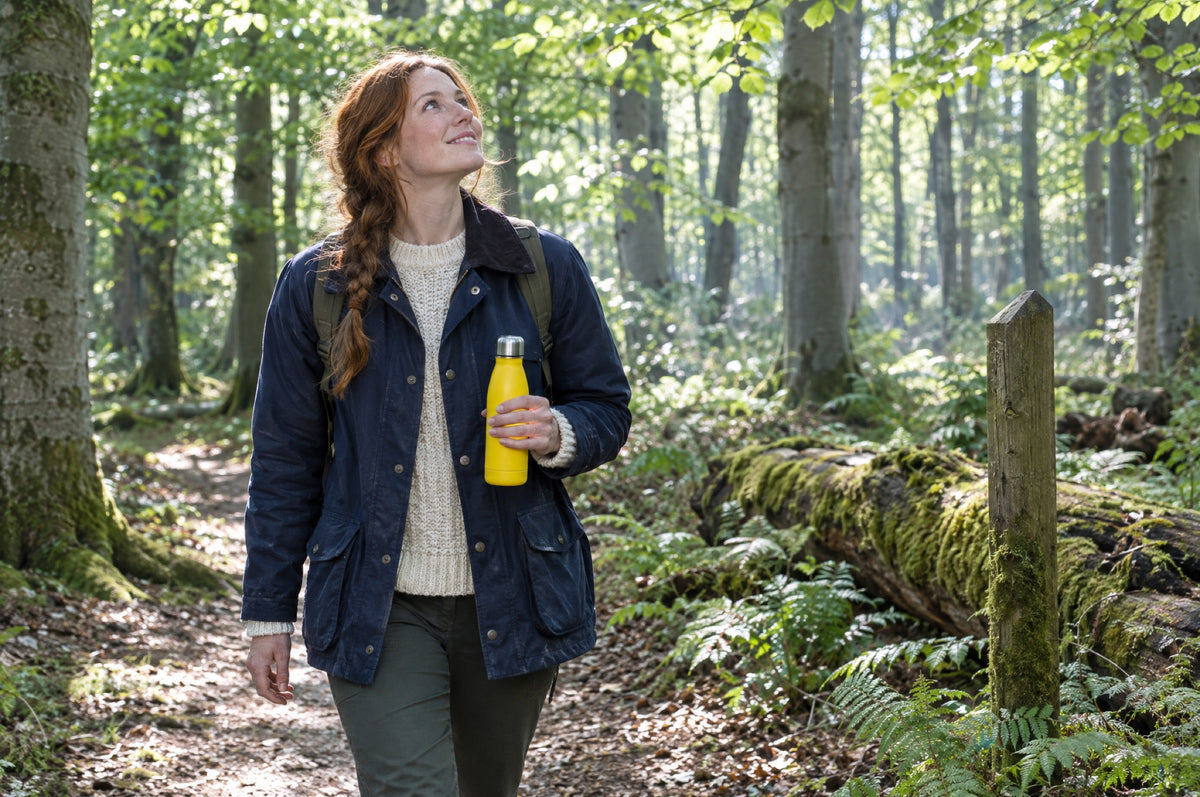 A young woman with a plait walks through a sunlit beech wood holding a yellow flask.
