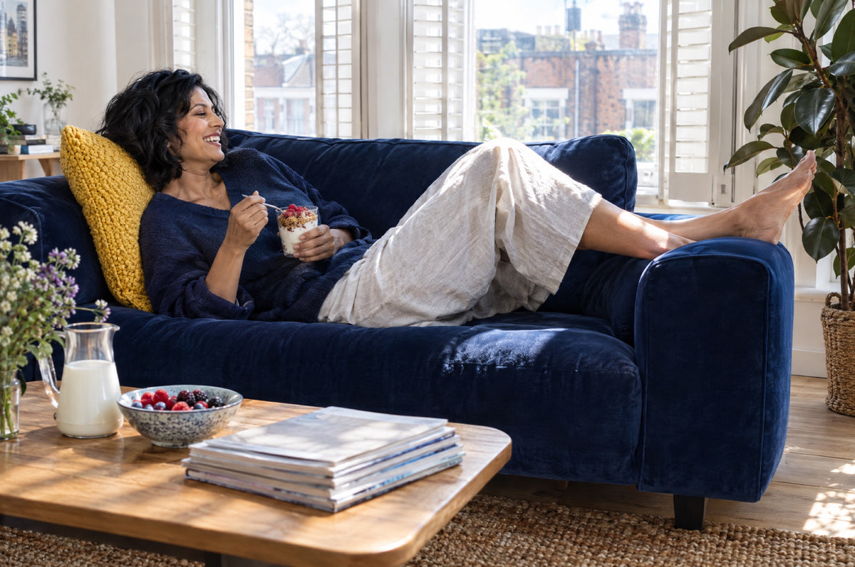 A woman laughs on a navy sofa with a yellow cushion holding a glass of yoghurt and raspberries in a sunlit flat.