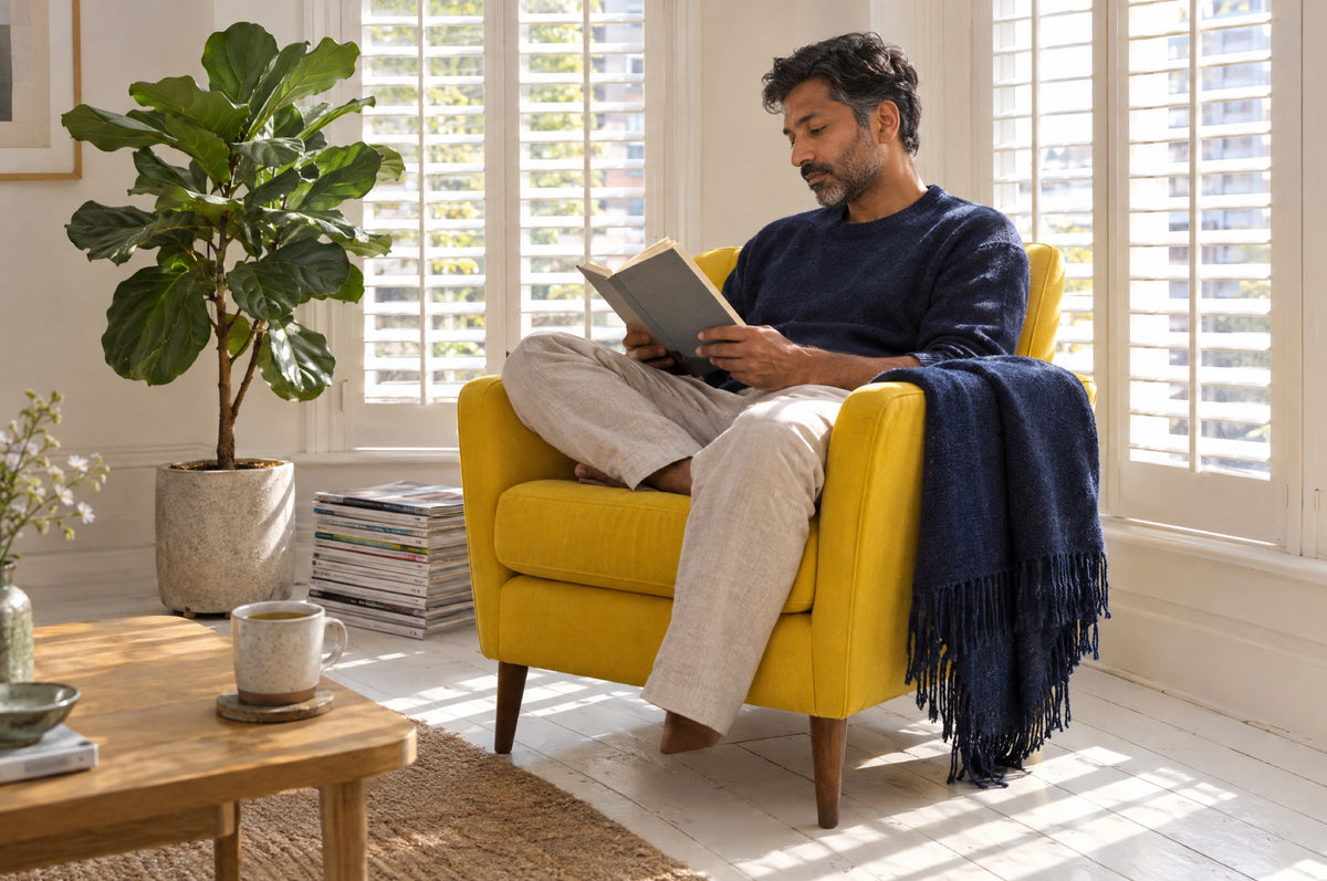 A man reads in a yellow armchair with a navy throw as afternoon light falls through shutters.