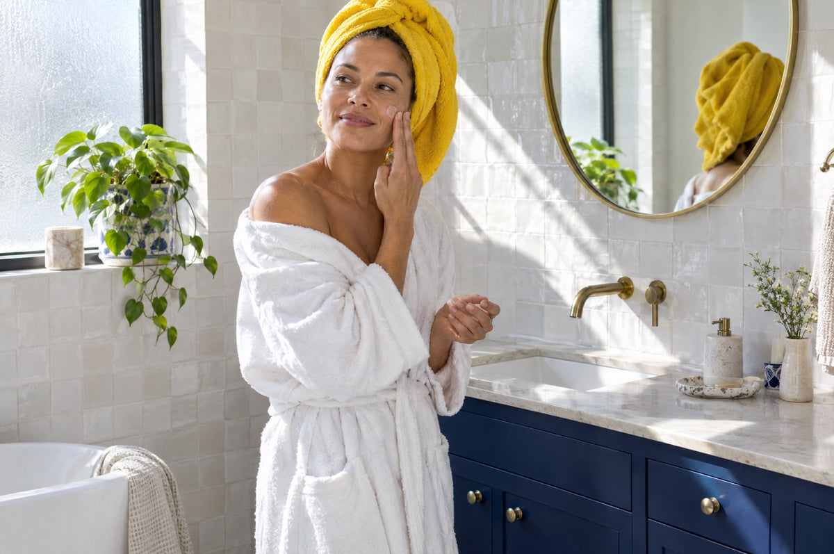 A woman with a yellow towel on her hair applies moisturiser in a bright bathroom with navy cabinets.