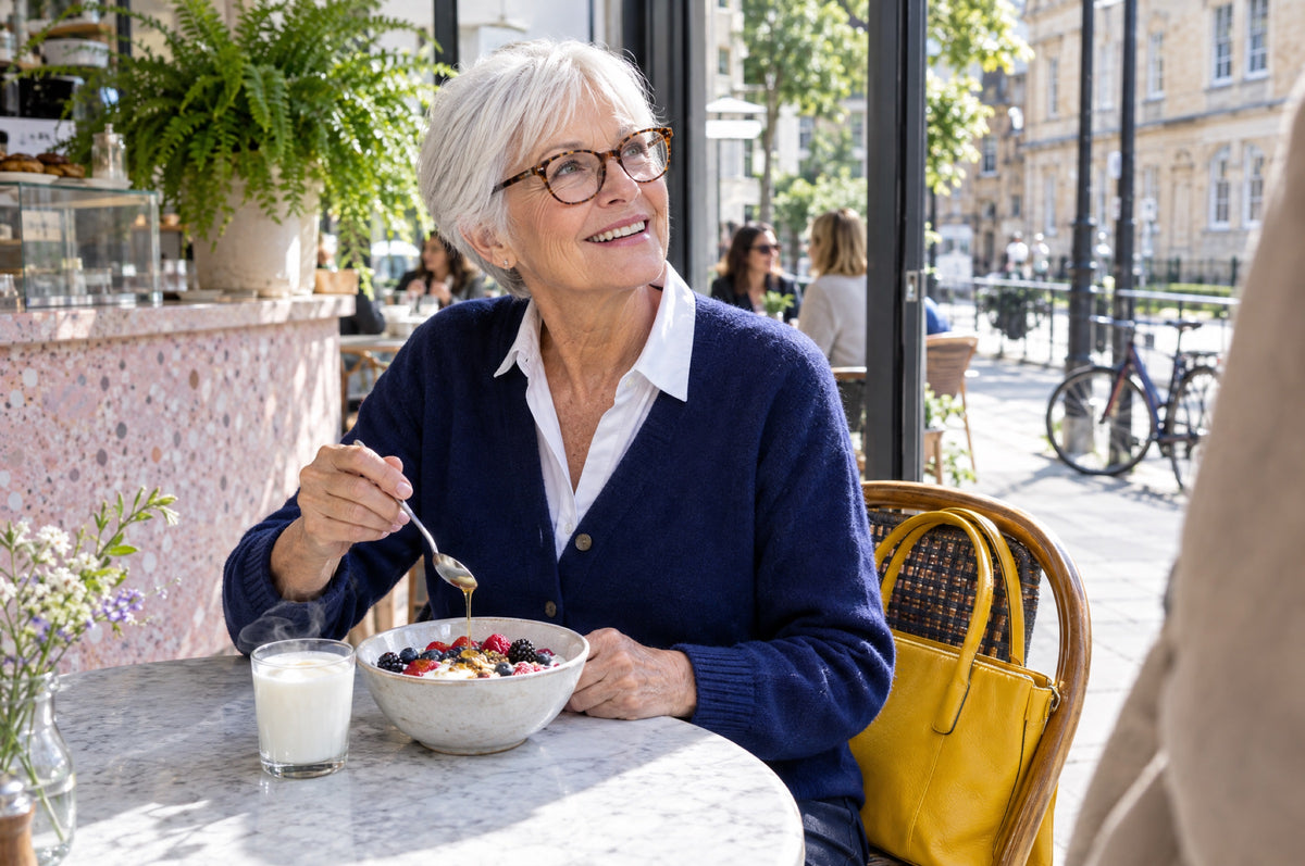 An older woman stirs honey into yoghurt and berries at a sunny café table with a yellow handbag on the chair beside her.