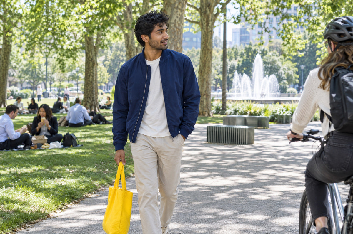 A young man walks through a sunny park carrying a yellow tote bag and wearing a navy jacket.