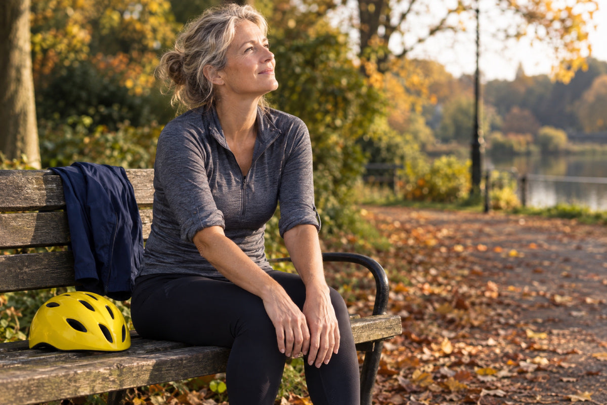 A woman in her fifties sits on a park bench in low autumn sun, sleeves rolled and face turned up to the light, hands on her knees, a plain yellow bike helmet beside her and a navy running jacket over the backrest.