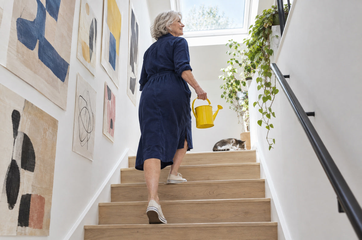 An older woman climbs a bright staircase carrying a yellow watering can towards plants on the landing.