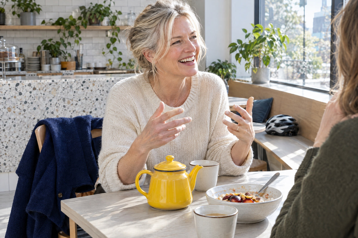 A woman laughs with a friend over tea from a yellow teapot in a bright café, a navy coat on her chair.