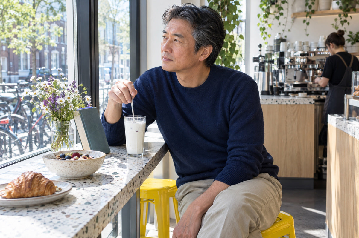 A man stirs a glass of kefir at a sunny café window counter, sitting on a yellow stool.