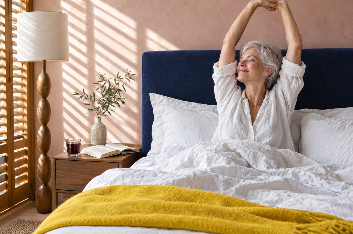 An older woman stretches in bed against a navy headboard with a yellow throw and a glass of cherry juice nearby.