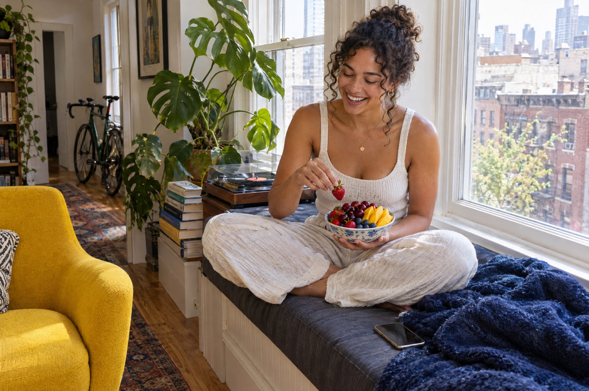 A young woman laughs on a window seat with a bowl of fresh berries and a yellow armchair in front of her.