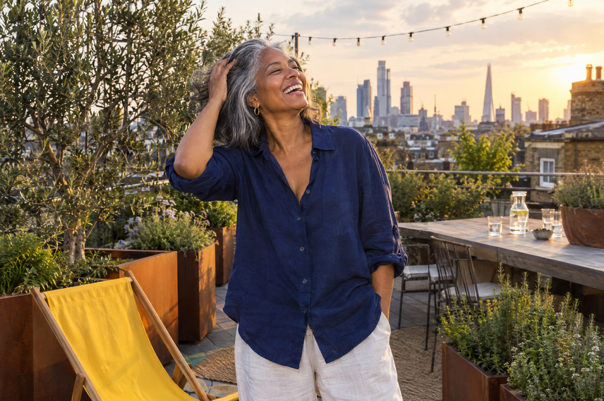 A woman in a navy shirt laughs on a rooftop terrace at sunset beside a yellow deck chair.