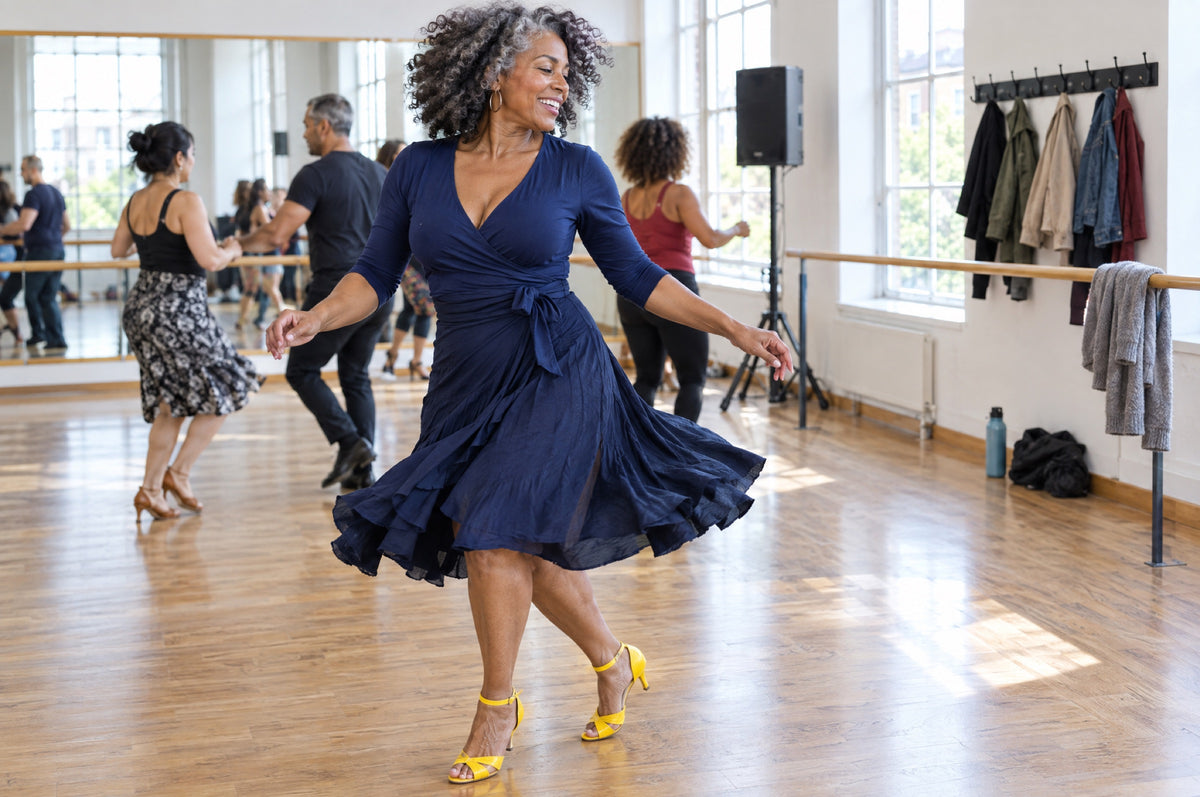 A woman in a navy top and yellow dance shoes spins mid-step in a sunlit dance studio.
