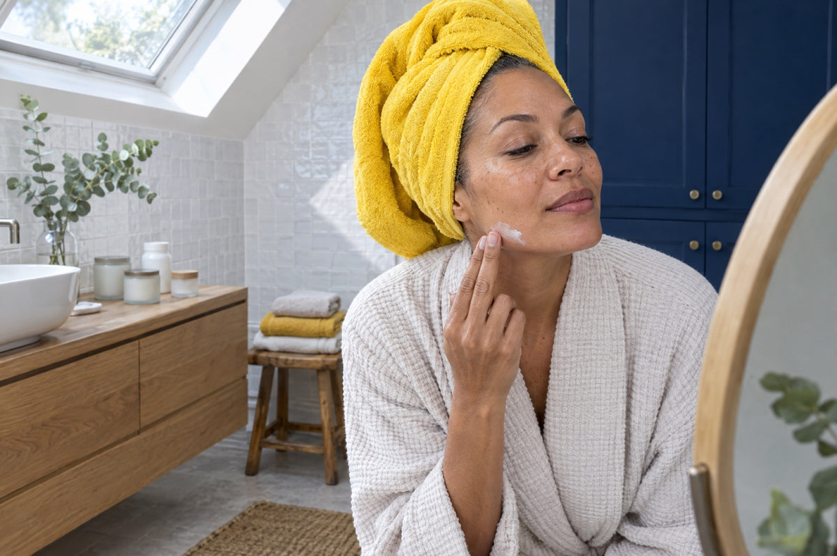 A woman with her hair in a yellow towel smooths moisturiser along her jaw in a bright tiled bathroom.