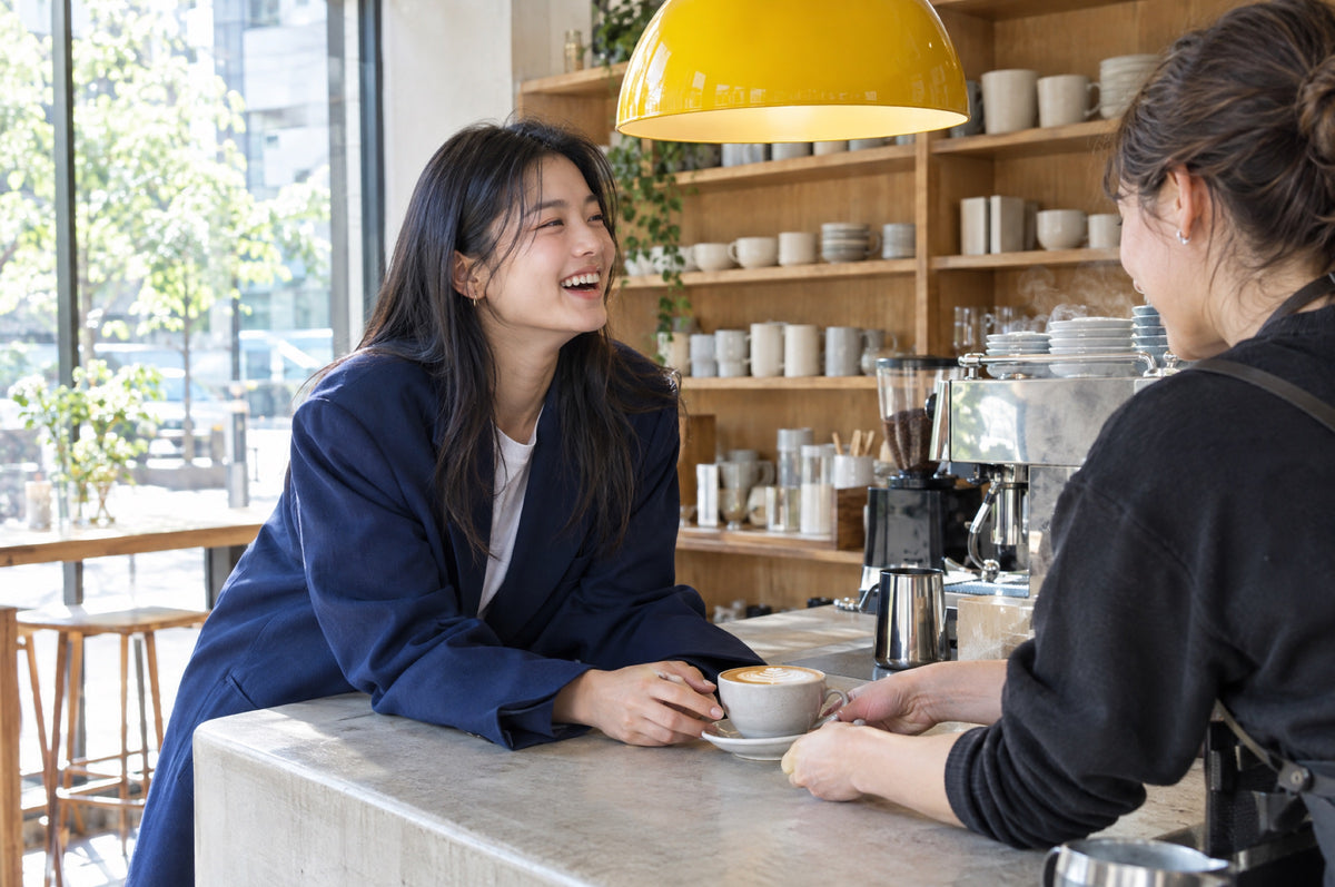 A young woman laughs with a barista as she collects a flat white under a yellow pendant lamp.