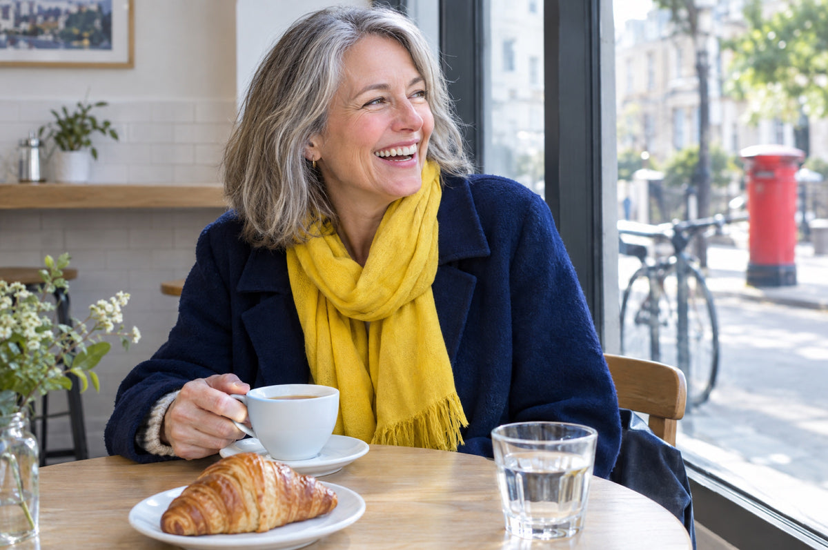 A woman with grey hair in a navy coat and yellow scarf laughs over a coffee at a café window table, a red postbox on the street outside.