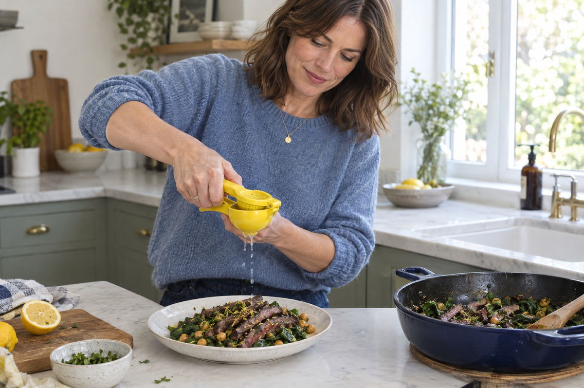 A woman in a blue jumper squeezes lemon with a yellow citrus press over a plate of steak strips, spinach and chickpeas, a navy cast-iron pan beside her.