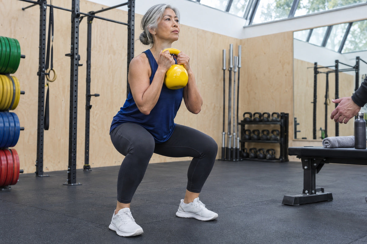 An older woman in a navy vest lifts a yellow kettlebell in a bright skylit gym.