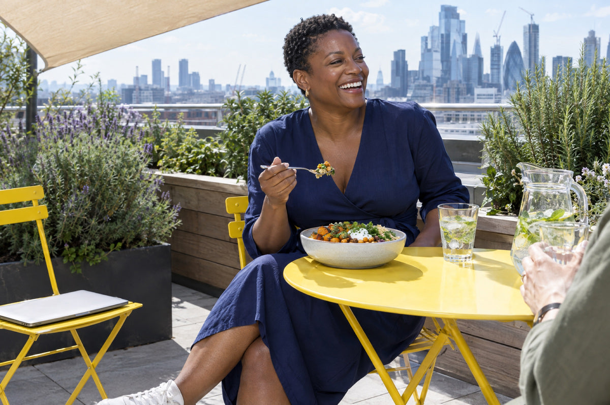 A woman in a navy dress laughs over a colourful lunch bowl on a rooftop terrace, seated on a yellow chair.