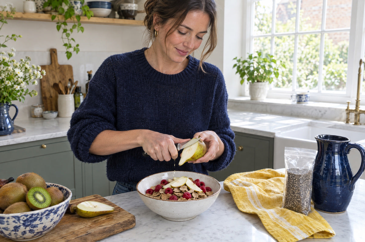 A woman in a navy jumper slices a pear over a bowl of bran flakes and raspberries at a kitchen counter, kiwis, a bag of chia seeds and a yellow tea towel beside her.