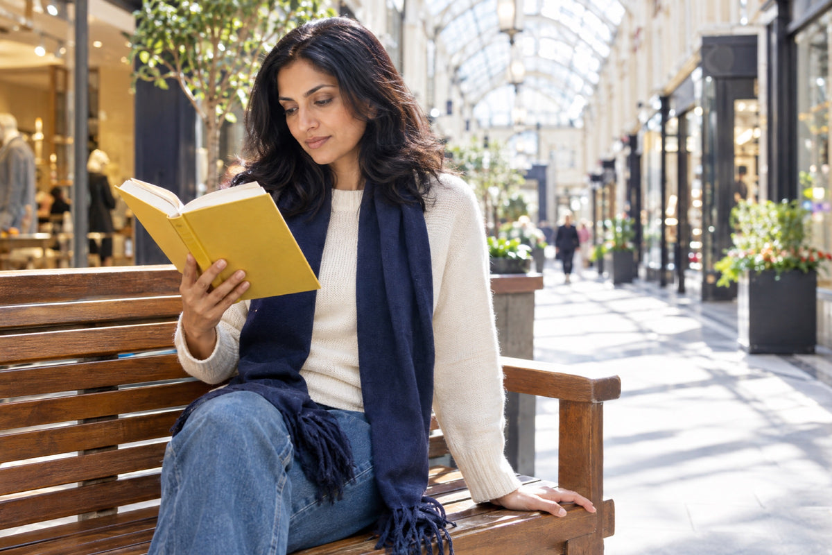 A woman in her early forties reads a yellow hardback on a wooden bench in a sunlit glass-roofed arcade, a navy scarf loose around her neck and one hand resting on the bench.