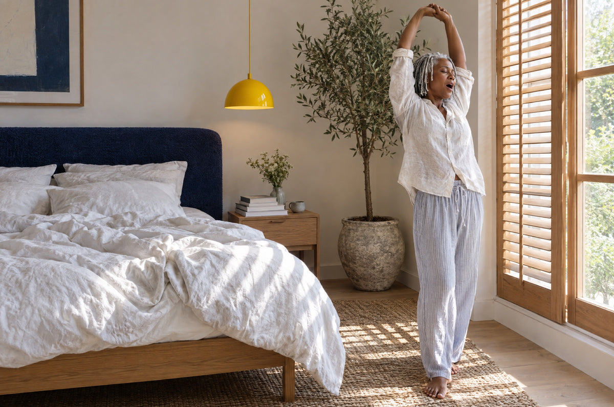 A woman stretches by a shuttered window in a bright bedroom with a yellow pendant lamp and a navy headboard.