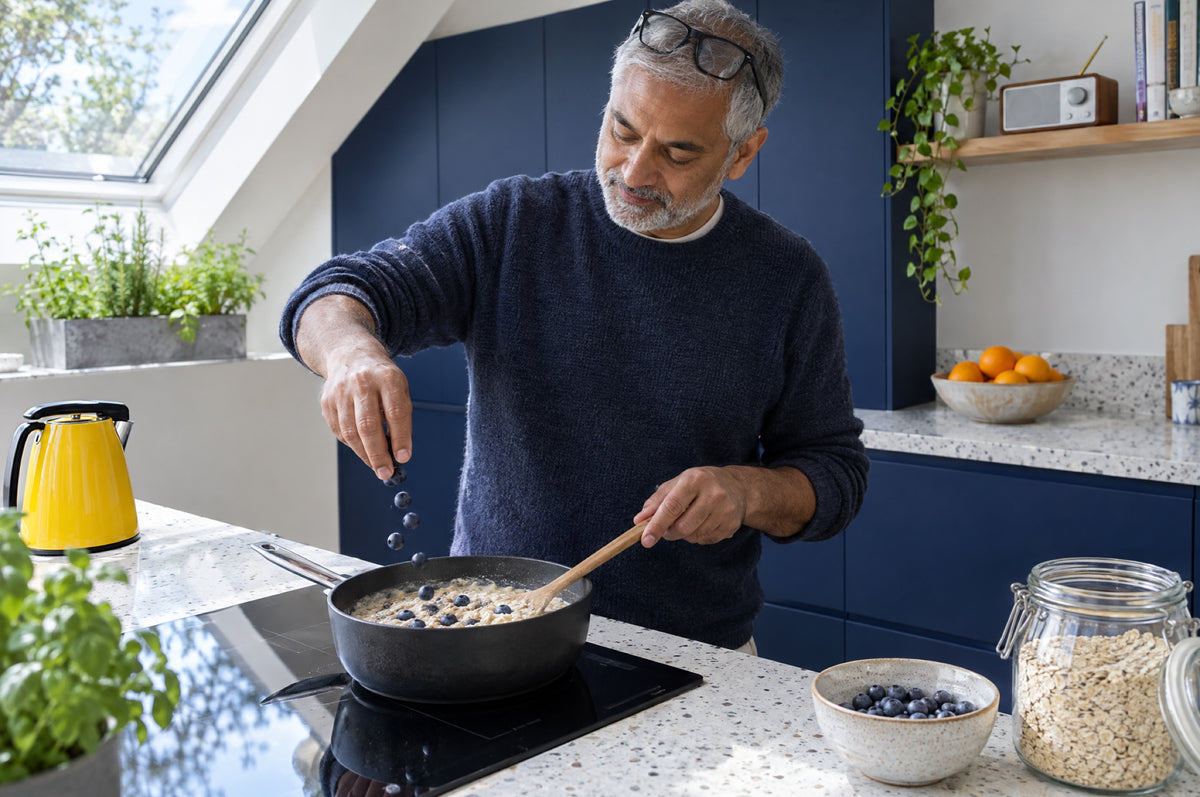 A grey-bearded man sprinkles blueberries onto porridge in a bright kitchen with a yellow kettle and navy cabinets.