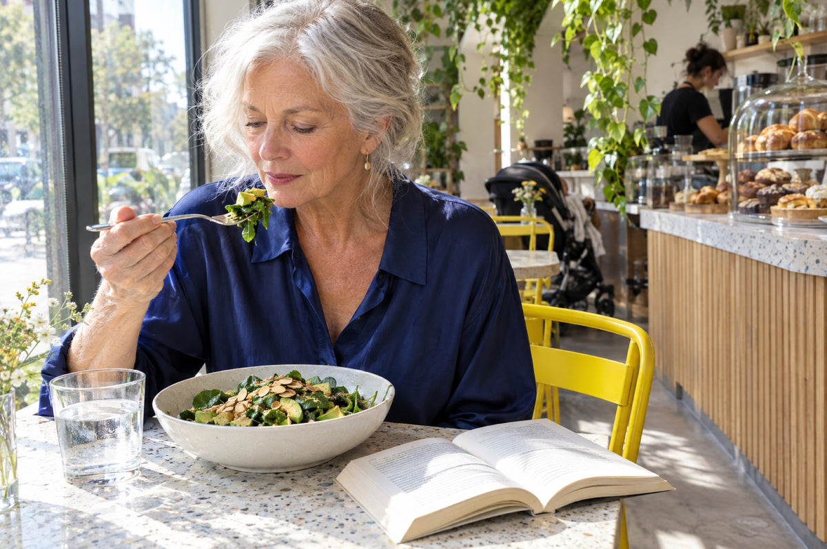 An older woman eats a green salad at a sunny café window, seated on a yellow chair.