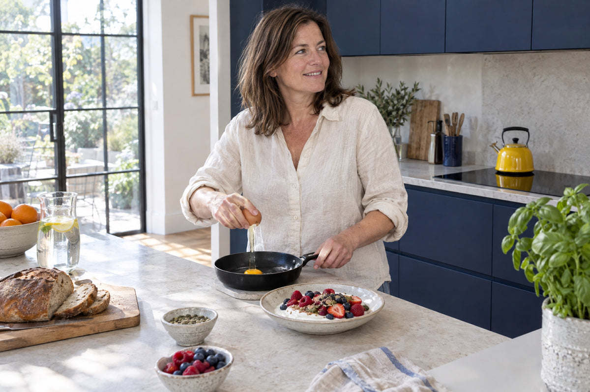 A woman cracks an egg into a pan in a bright kitchen with navy cabinets, a yellow kettle and a bowl of yoghurt and berries on the counter.