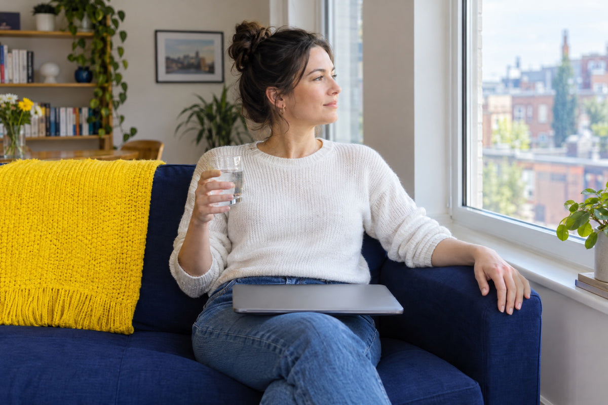 A woman in her thirties sits on a navy sofa in a bright flat with a closed laptop on her knees, a glass of water in one hand, a yellow throw over the sofa back, looking out of a tall window