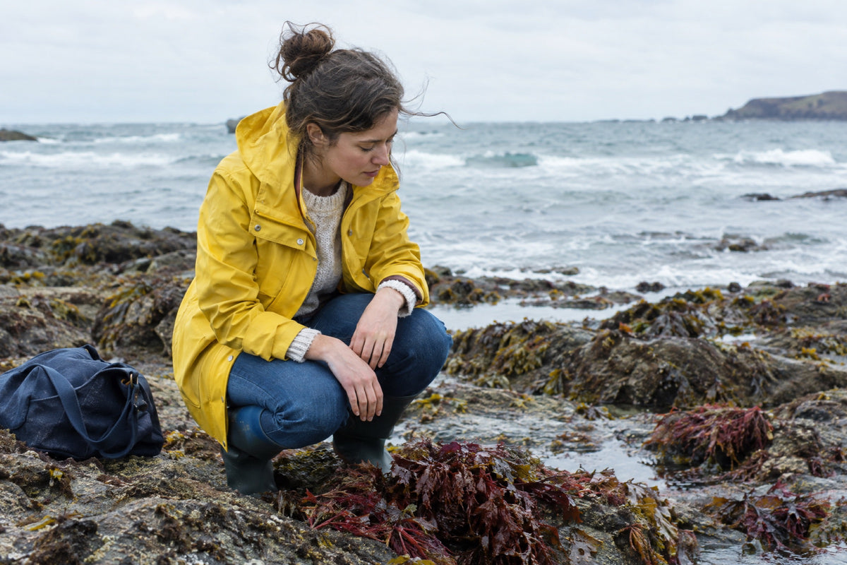 A British woman in a yellow raincoat on a windswept Cornish shoreline at low tide, crouching to look at red seaweed on the rocks, a navy canvas bag over her shoulder