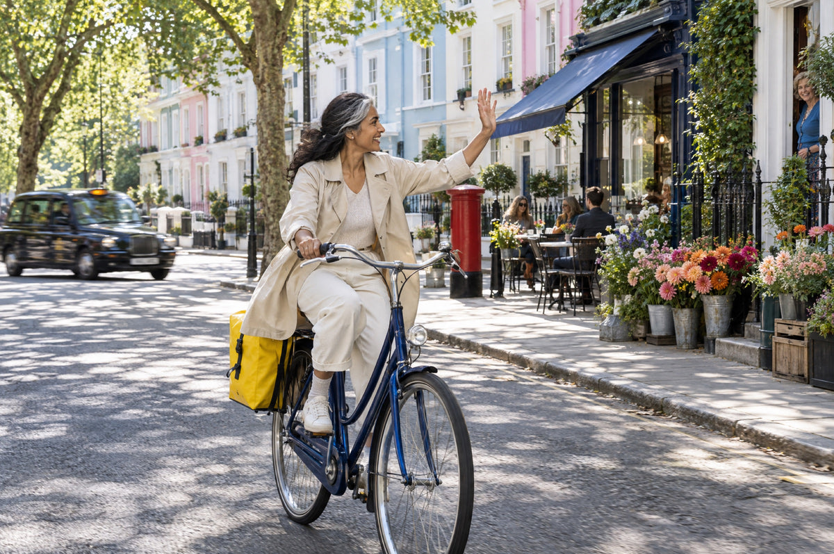 A woman rides a navy bicycle with a yellow pannier down a sunny street of pastel houses.