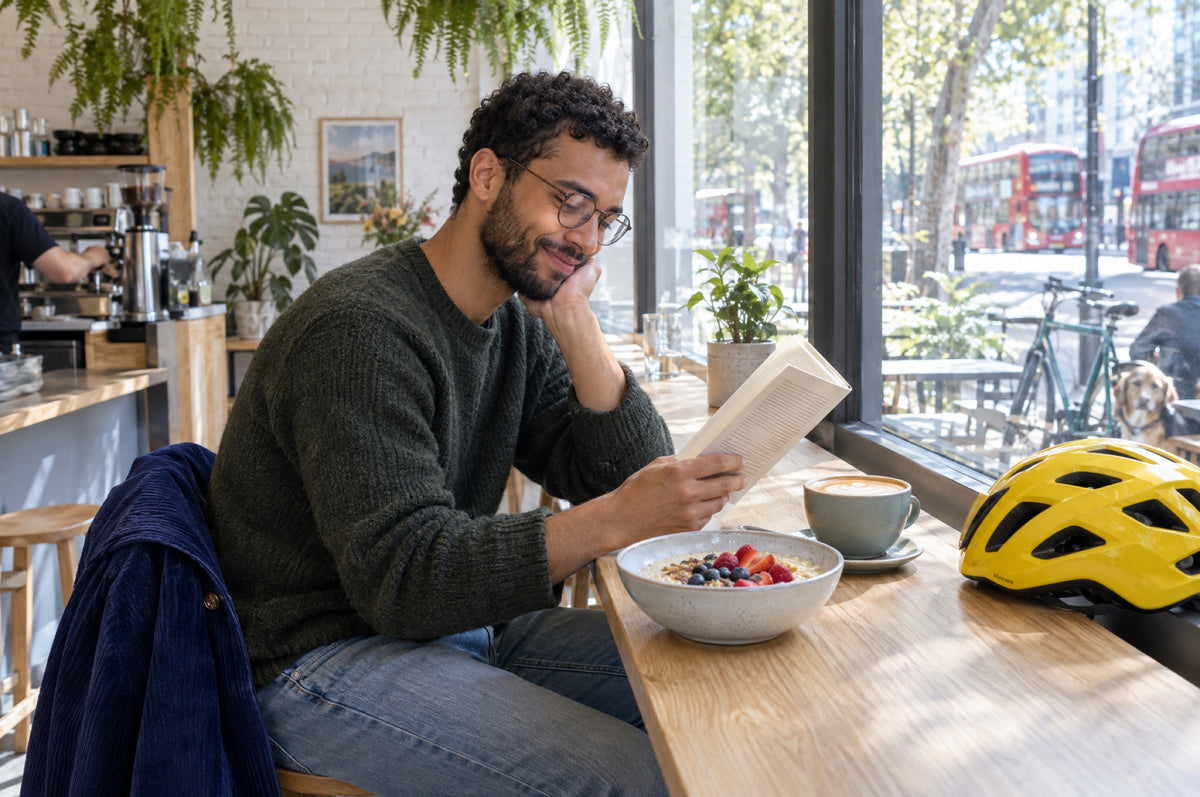 A man reads at a sunny café window beside a yellow cycling helmet, his navy jacket on the stool.