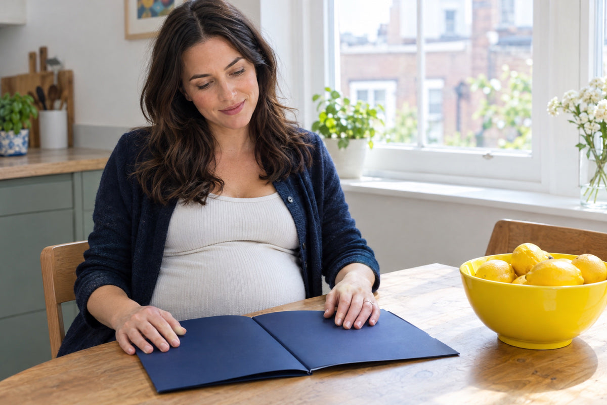 A pregnant woman in her thirties sits at a sunlit kitchen table, one hand resting on a closed navy folder and the other on her bump, a yellow bowl of lemons beside her and a bright window behind.
