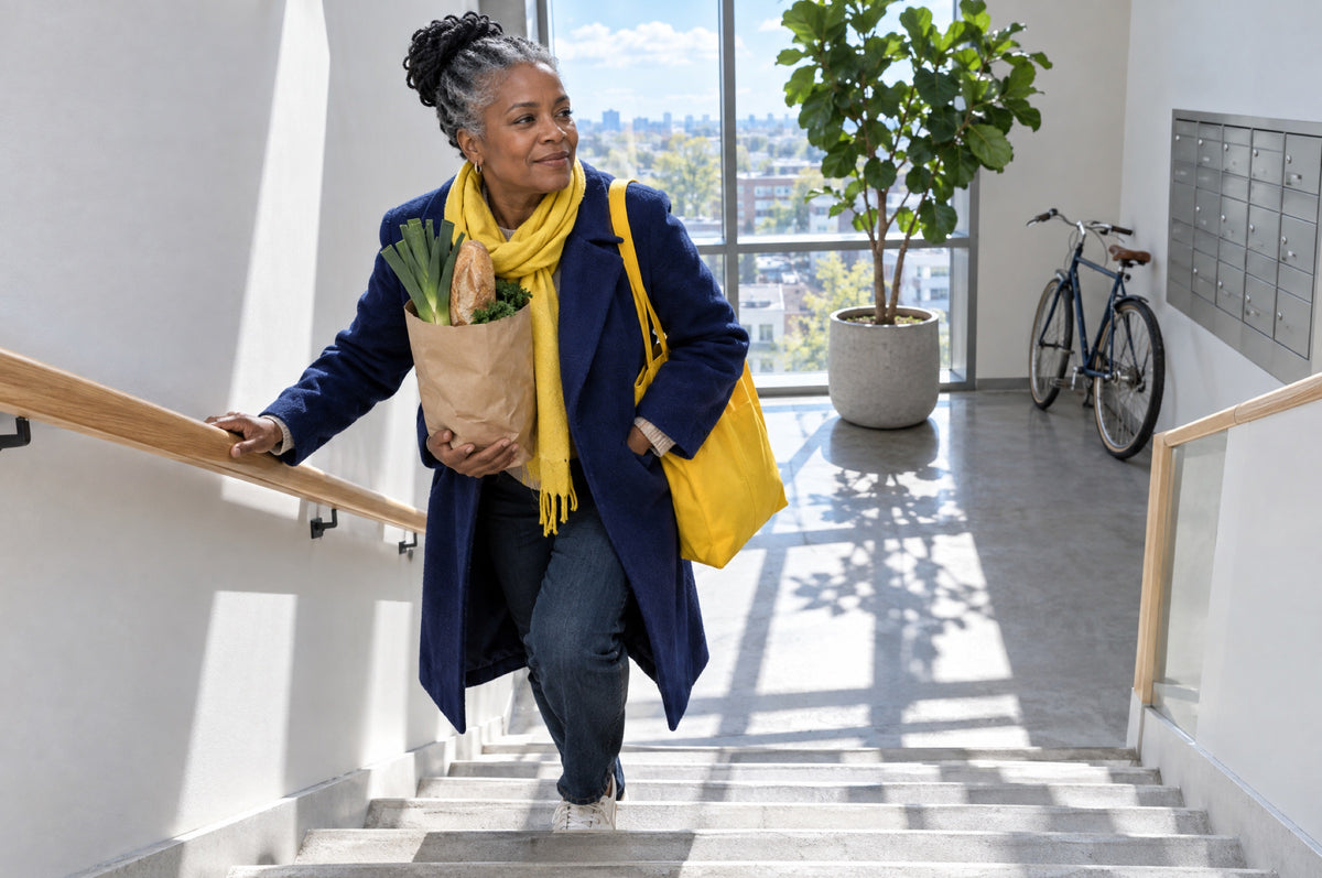 An older woman carries groceries up a bright modern stairwell wearing a navy coat and a yellow bag.