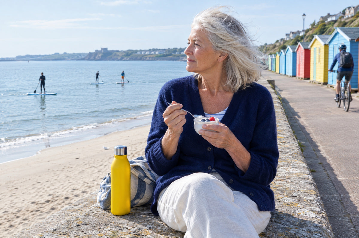 An older woman eats yoghurt on a sunny sea wall with a yellow flask beside her.