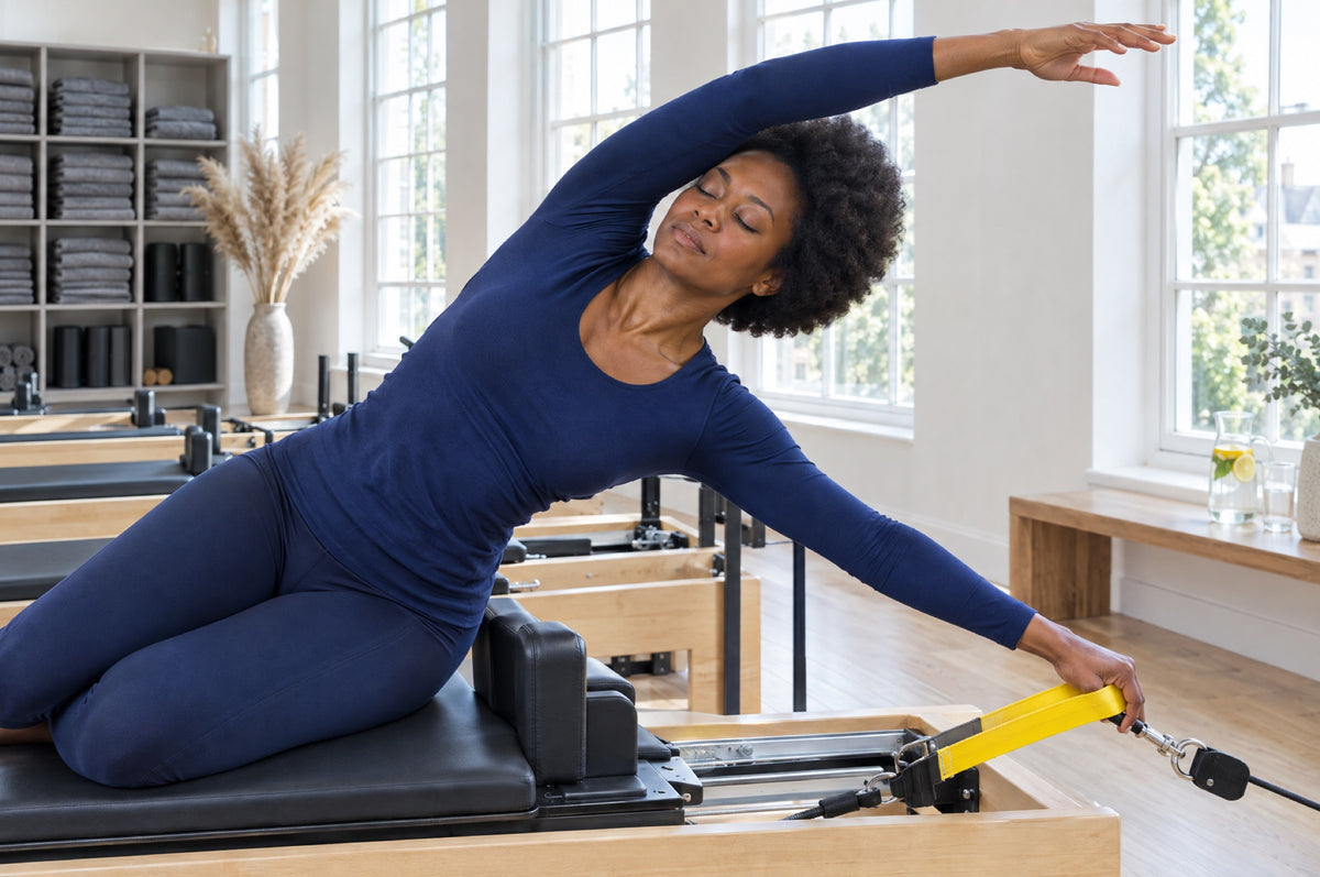 A woman in a navy top stretches sideways on a reformer holding a yellow strap in a sunlit pilates studio.