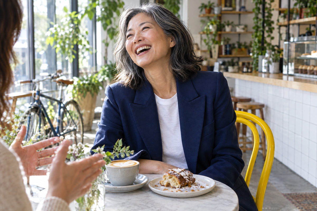 A woman in a navy jacket laughs at a café table while sitting on a yellow chair.