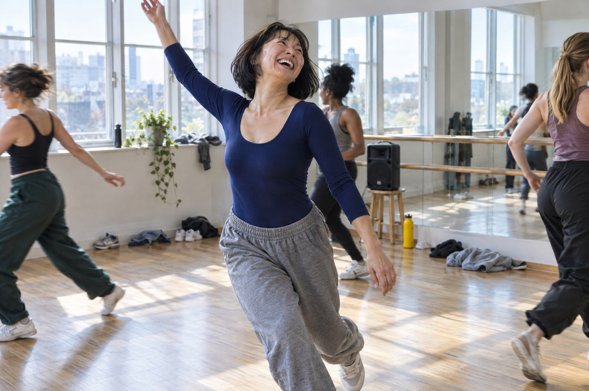 A woman in a navy leotard laughs mid-step in a sunlit dance studio with a yellow water bottle by the mirror.