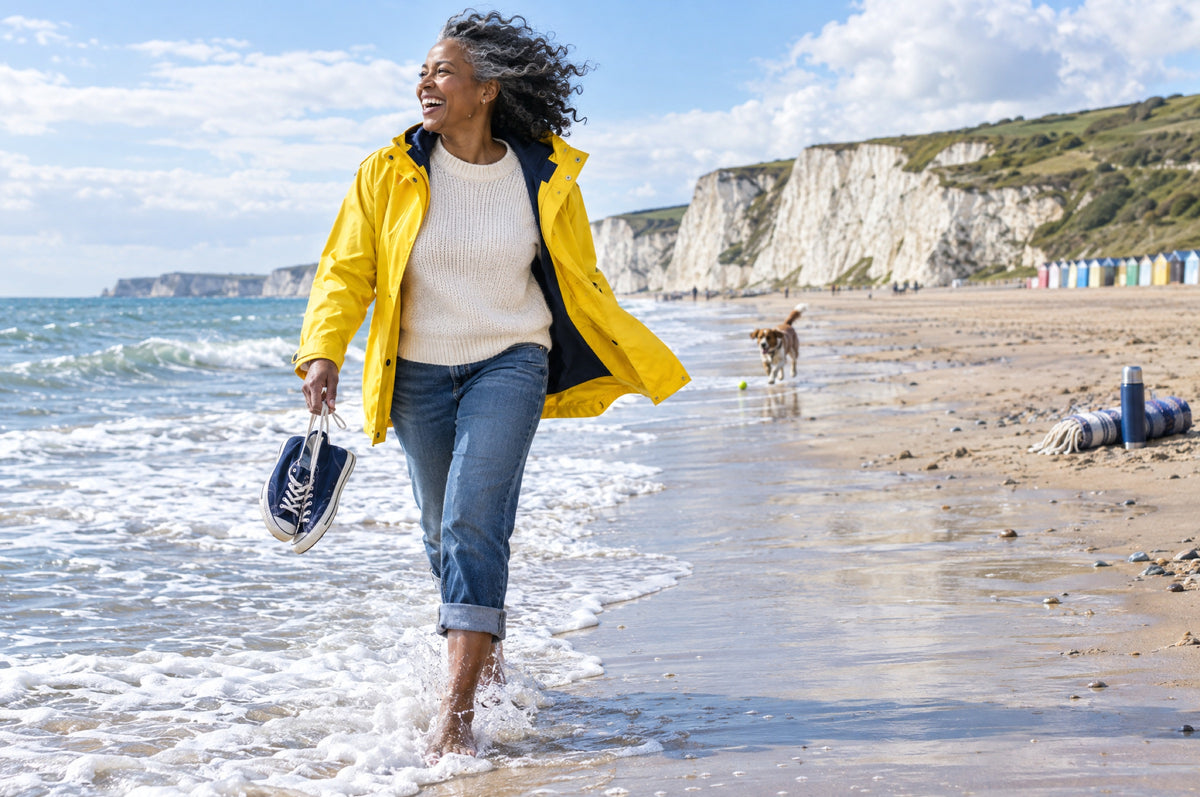 A woman in a yellow jacket laughs as a wave rushes over her bare feet on a wide beach.