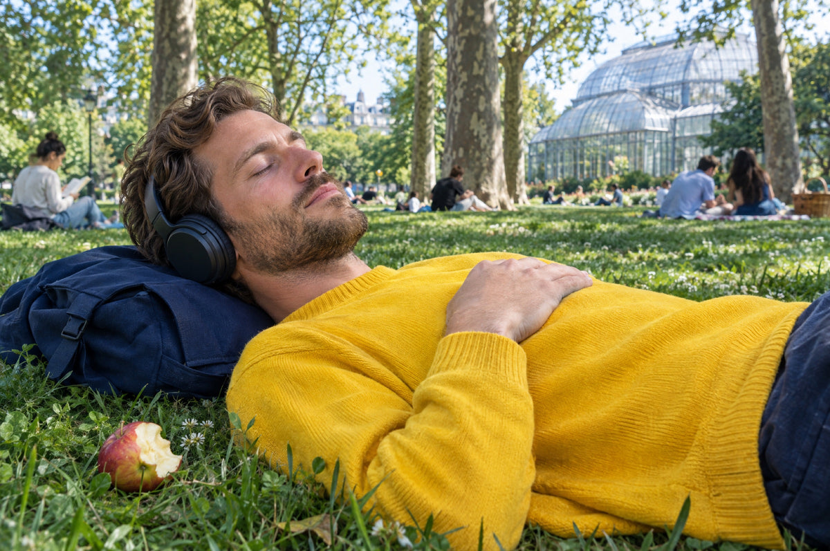 A man in a yellow jumper lies on park grass with headphones on, resting his head on a navy backpack.