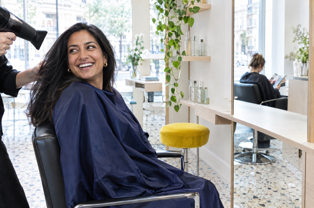 A woman laughs in a salon chair while her hair is blow-dried, a yellow stool at the next station.