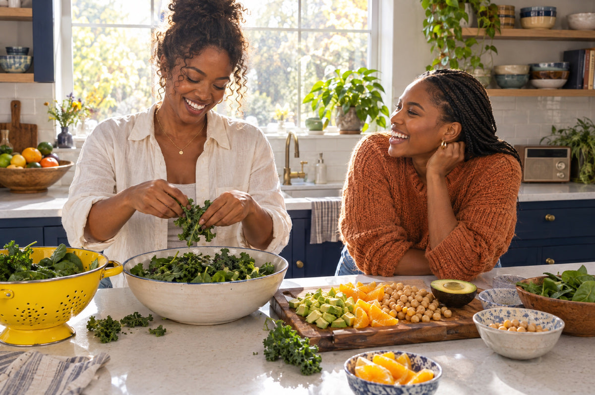 Two women laugh while tearing leafy greens into a bowl in a kitchen with navy cabinets and a yellow colander.