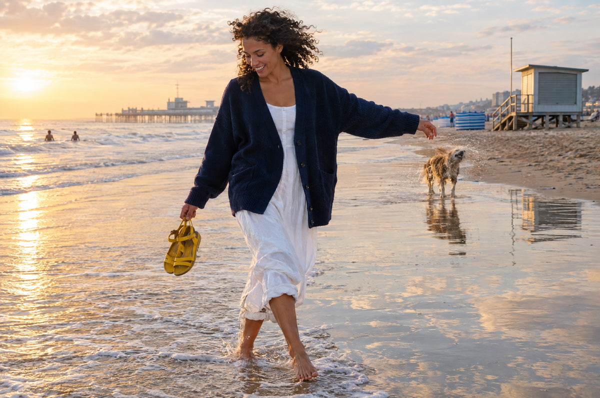 A woman in a navy cardigan paddles along the shoreline at golden hour holding yellow sandals.