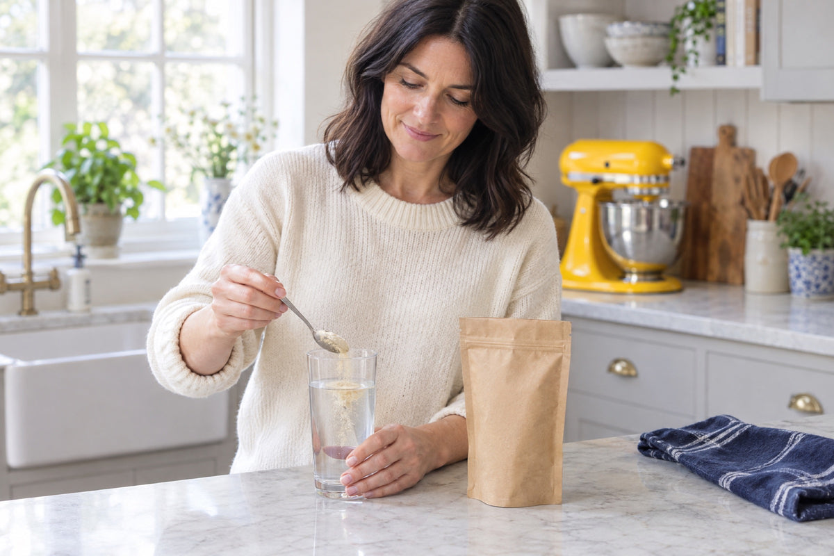 A dark-haired woman in a cream jumper stirs a spoonful of pale powder into a glass of water at a marble worktop beside a yellow stand mixer