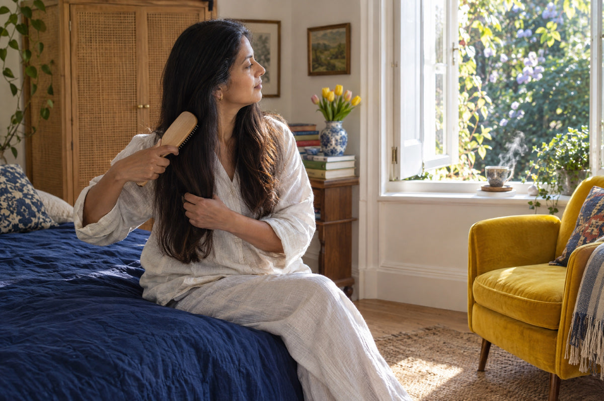 A woman brushes her long hair while sitting on a navy bedspread by an open bedroom window.