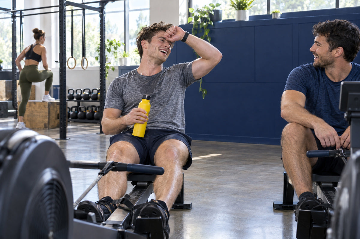 A young man laughs on a rowing machine in a bright gym holding a yellow water bottle.