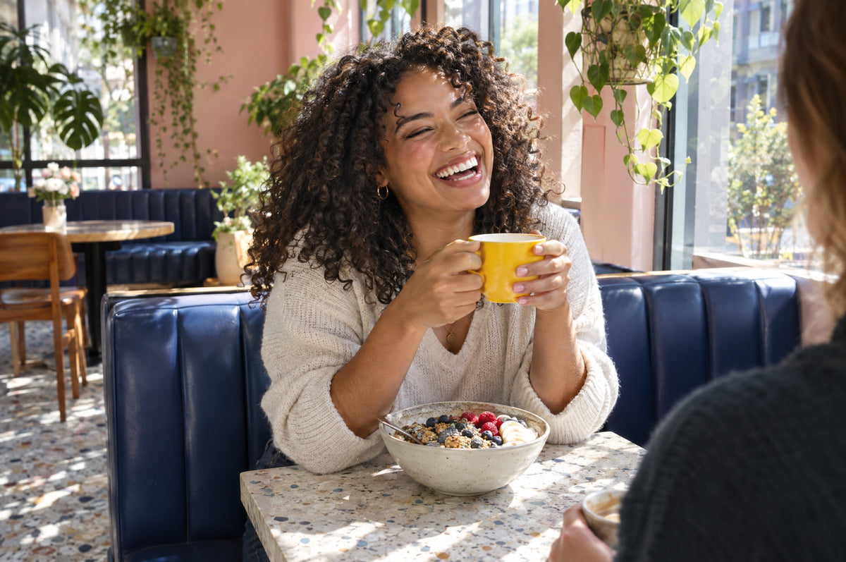A young woman laughs on a navy banquette in a sunny café holding a yellow cup.