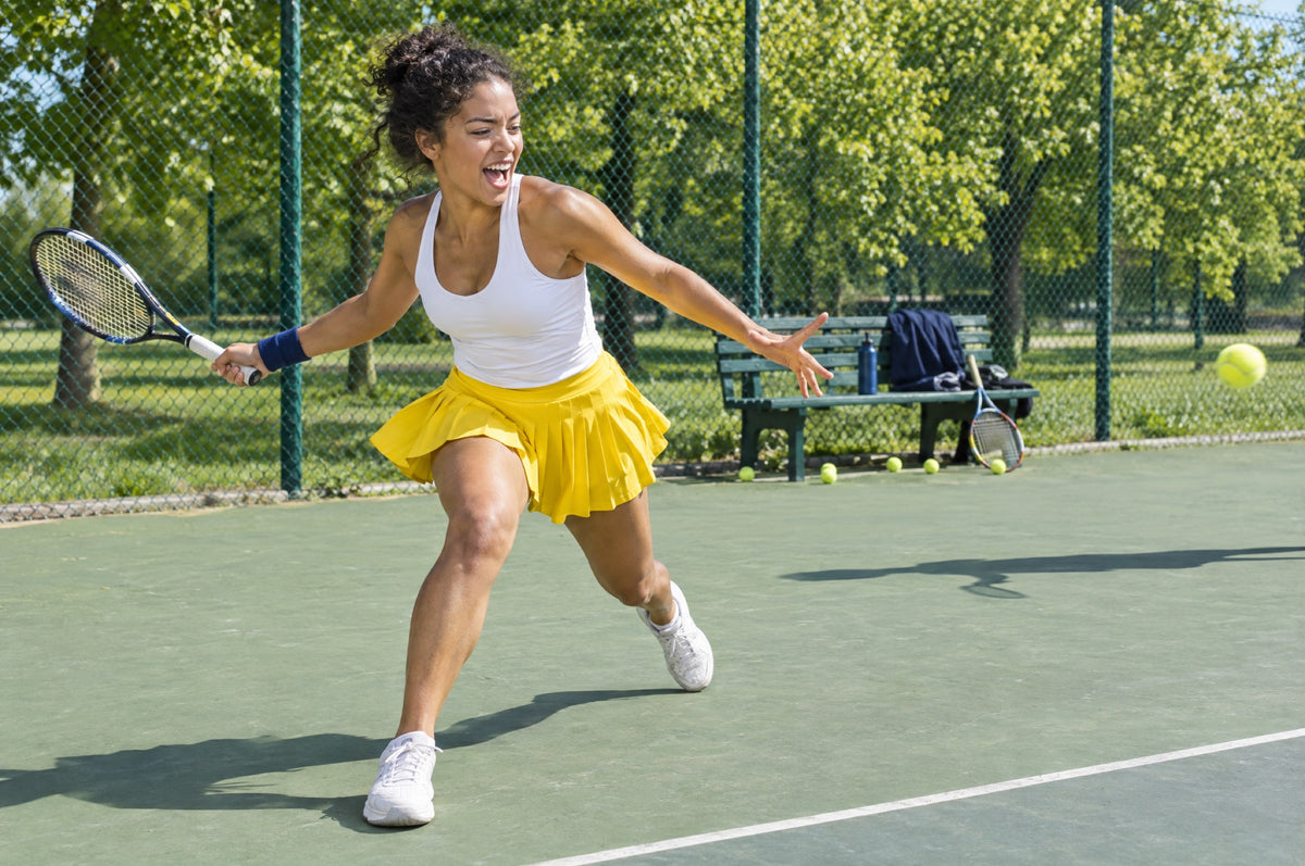 A young woman in a yellow skirt lunges for a tennis shot on a sunny outdoor court.