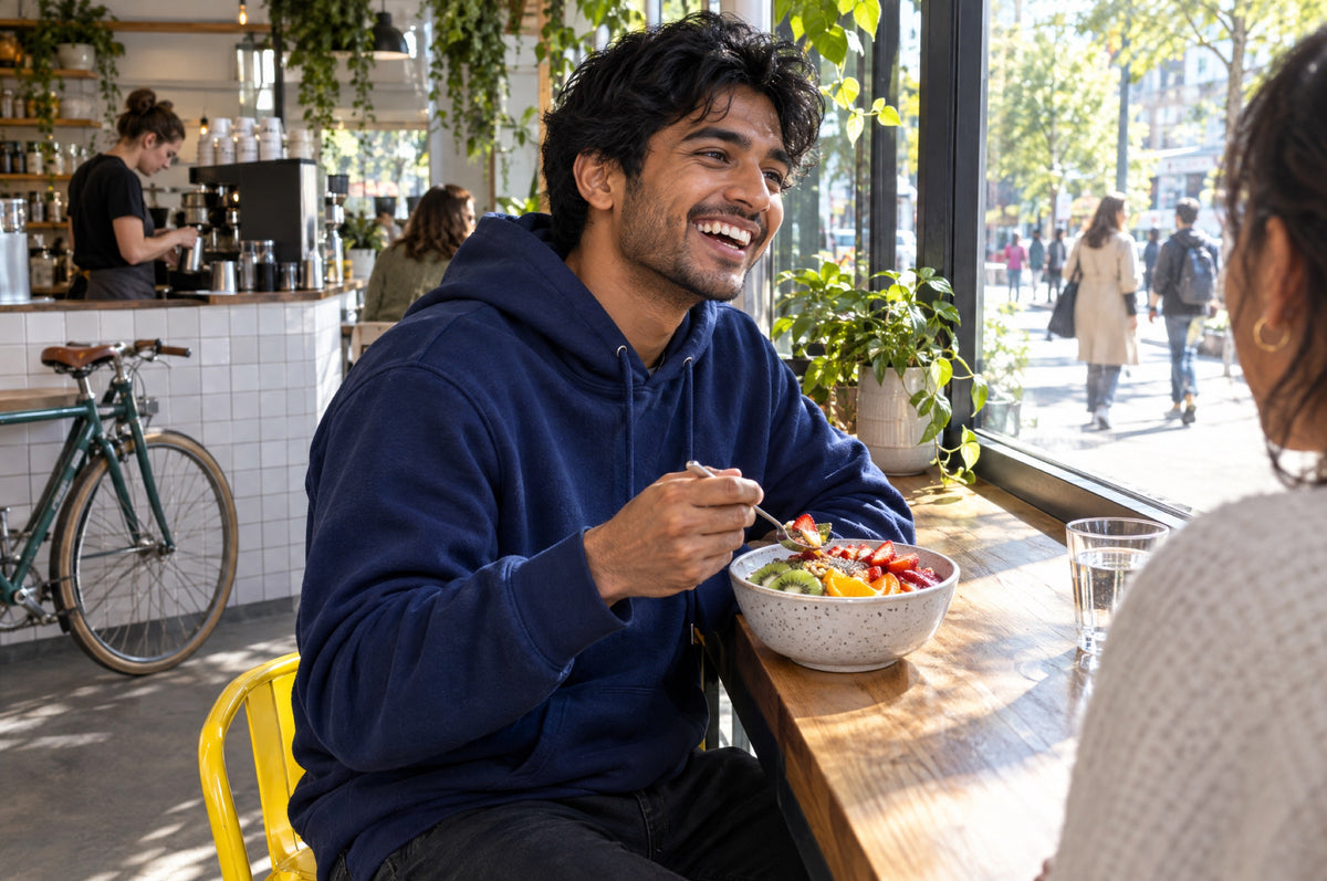 A young man eats a colourful fruit bowl at a café window while seated on a yellow stool.