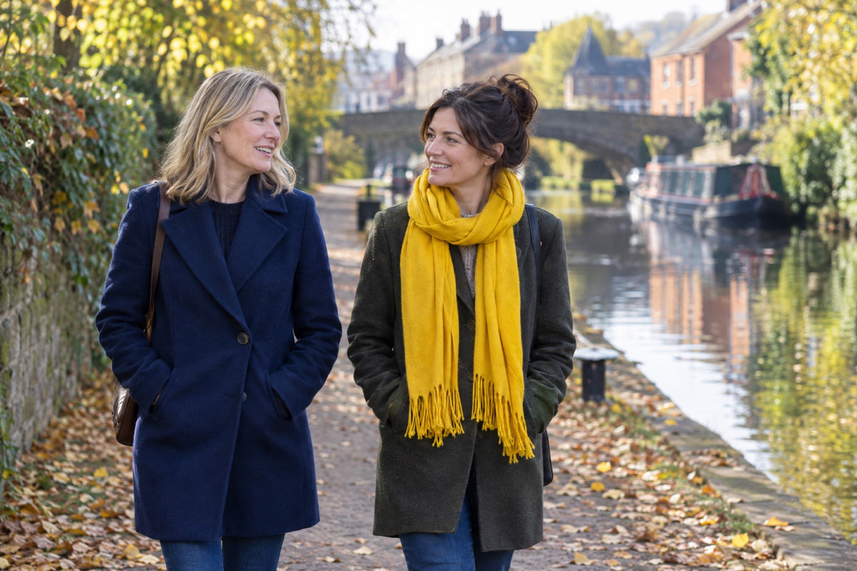 Two women in their forties walk along a canal towpath on a bright autumn morning, one in a navy wool coat and one wearing a yellow scarf, both with their hands in their coat pockets.